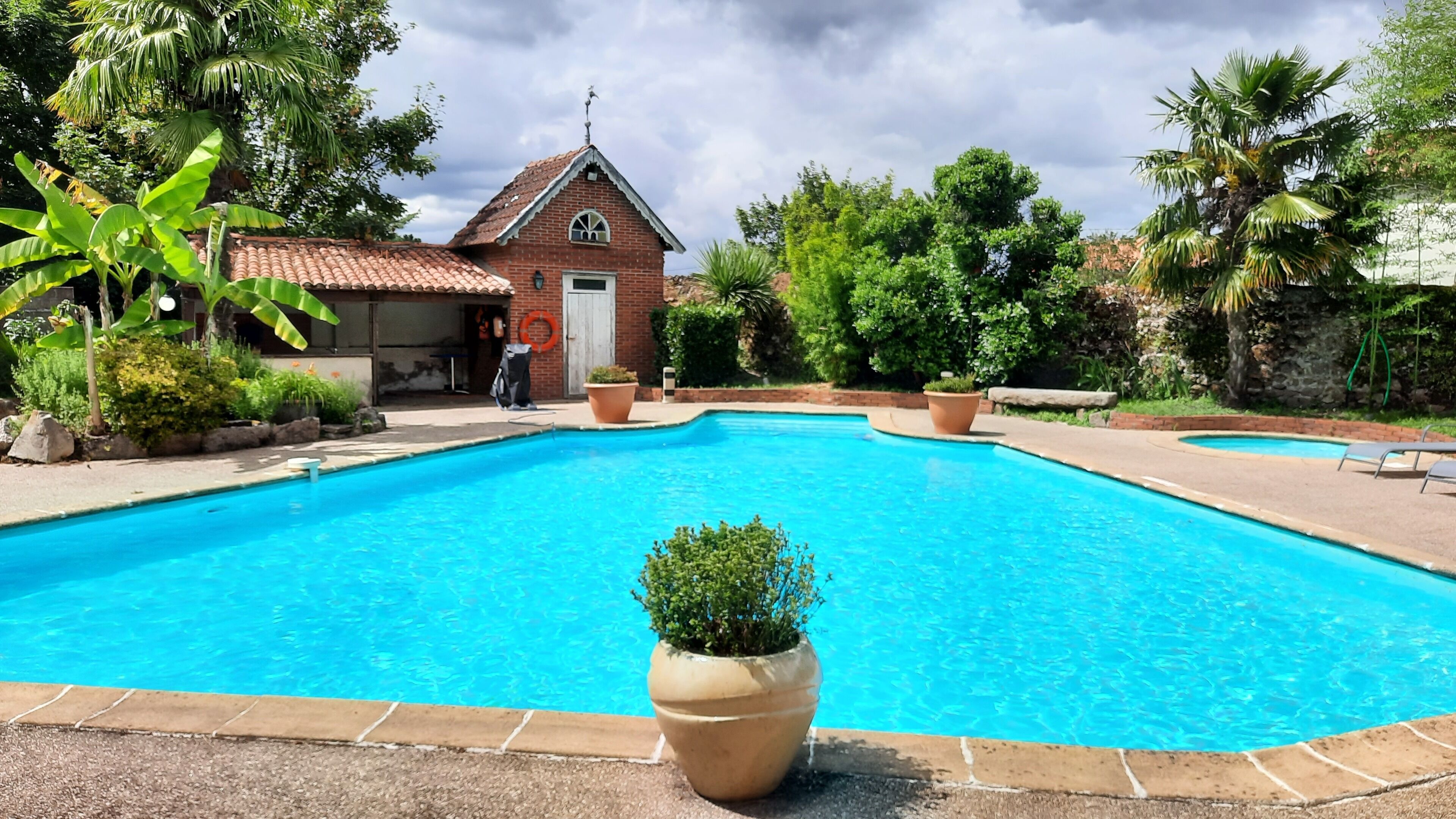 Una piscina rectangular con solárium alicatado, rodeada de exuberante vegetación y un pequeño edificio de ladrillo con tejado de tejas.