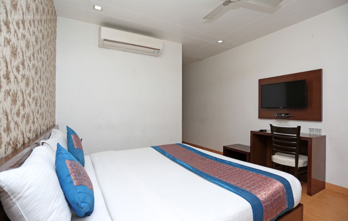 A hotel room with a bed, a desk, a chair, a TV mounted on the wall, and an air conditioning unit.