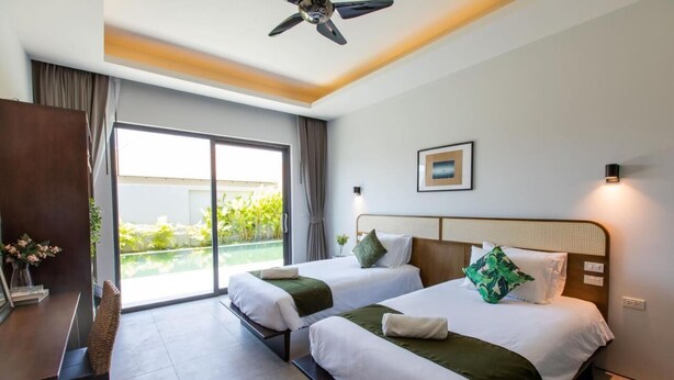 Habitació d'hotel amb dos llits, un escriptori i una cadira. Hi ha un ventilador de sostre, una finestra amb cortines i vista a vegetació a l'exterior.