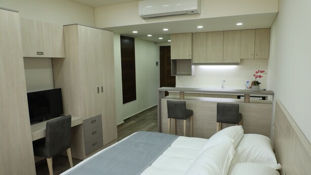 A hotel room with a bed, a desk, a chair, a kitchenette, and a TV. - byblos palms hotel (Blat)