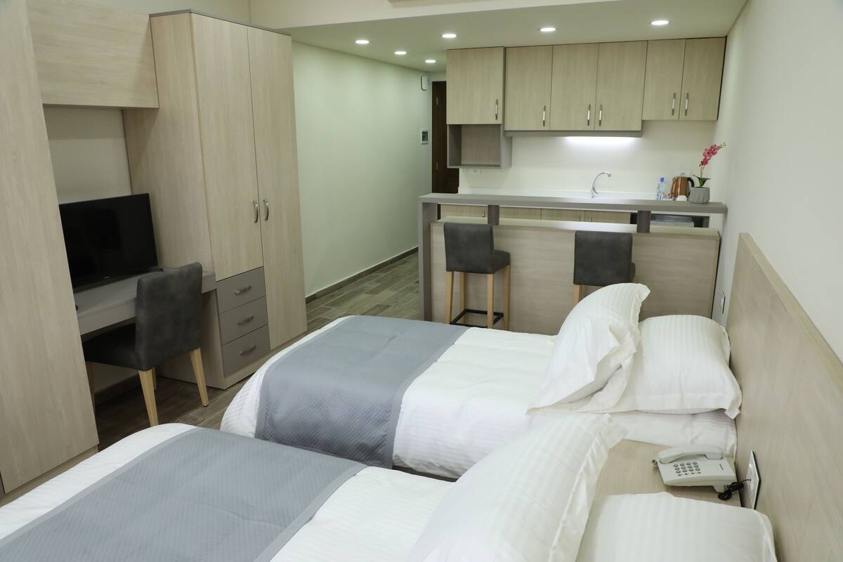 A hotel room with two beds, a desk with a chair, a TV, and a kitchenette.