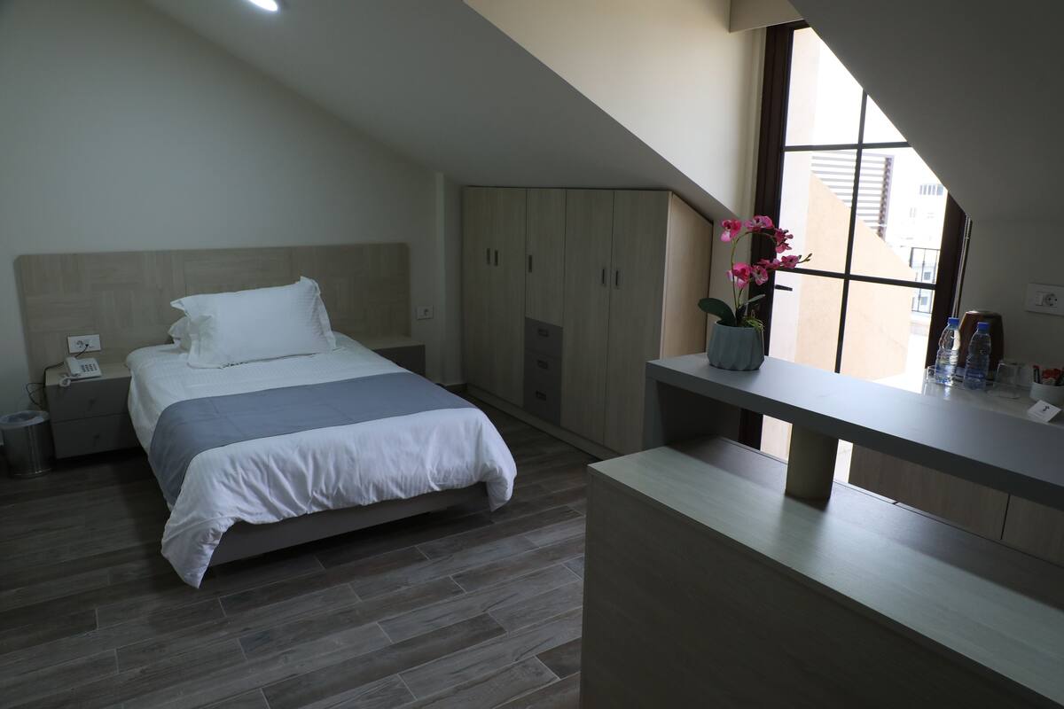 A bedroom with a bed, a nightstand, a wardrobe, a window, and a bedside table with a vase of flowers.