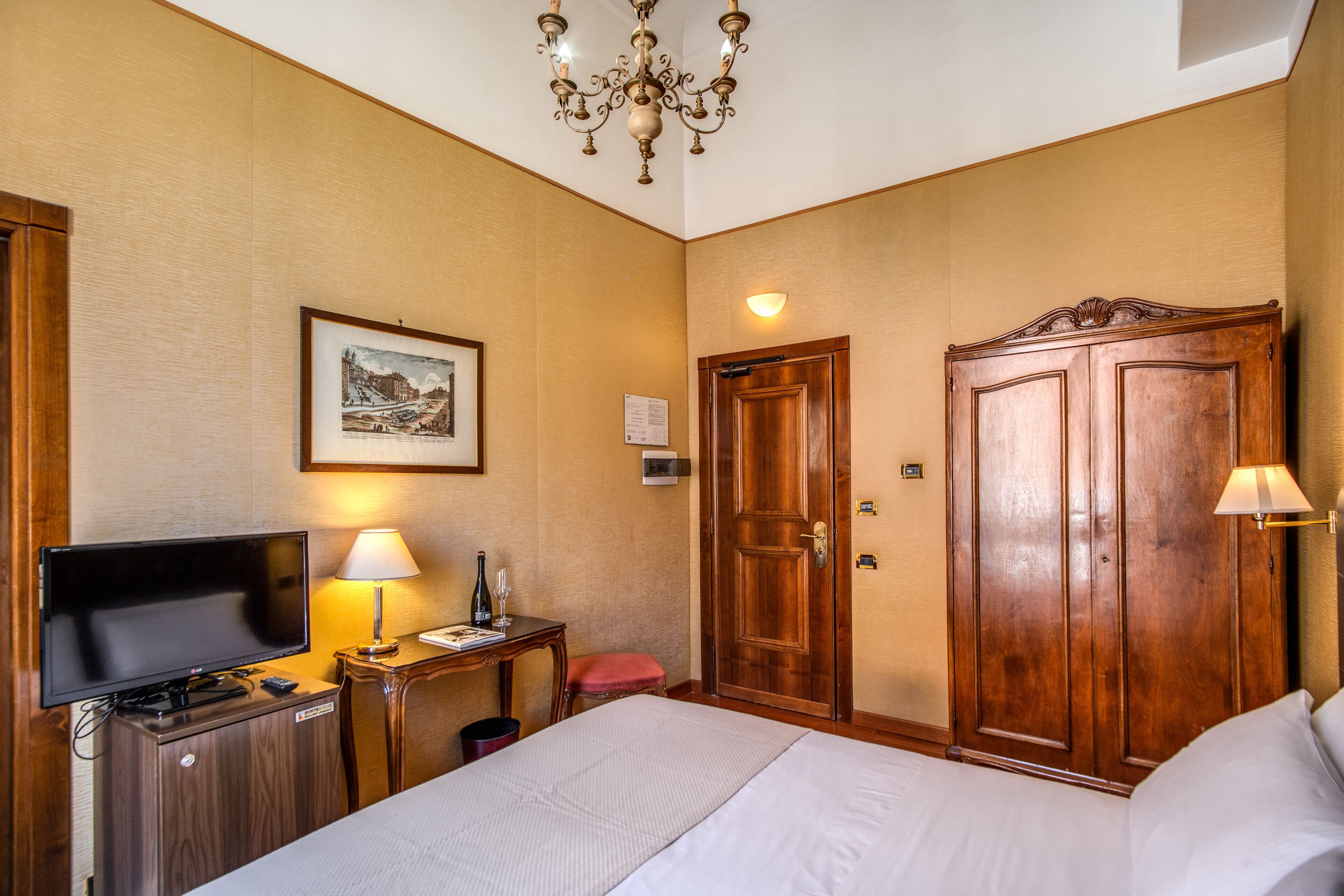 Habitación de hotel con cama, televisión, escritorio con lámpara, armario de madera y candelabro.