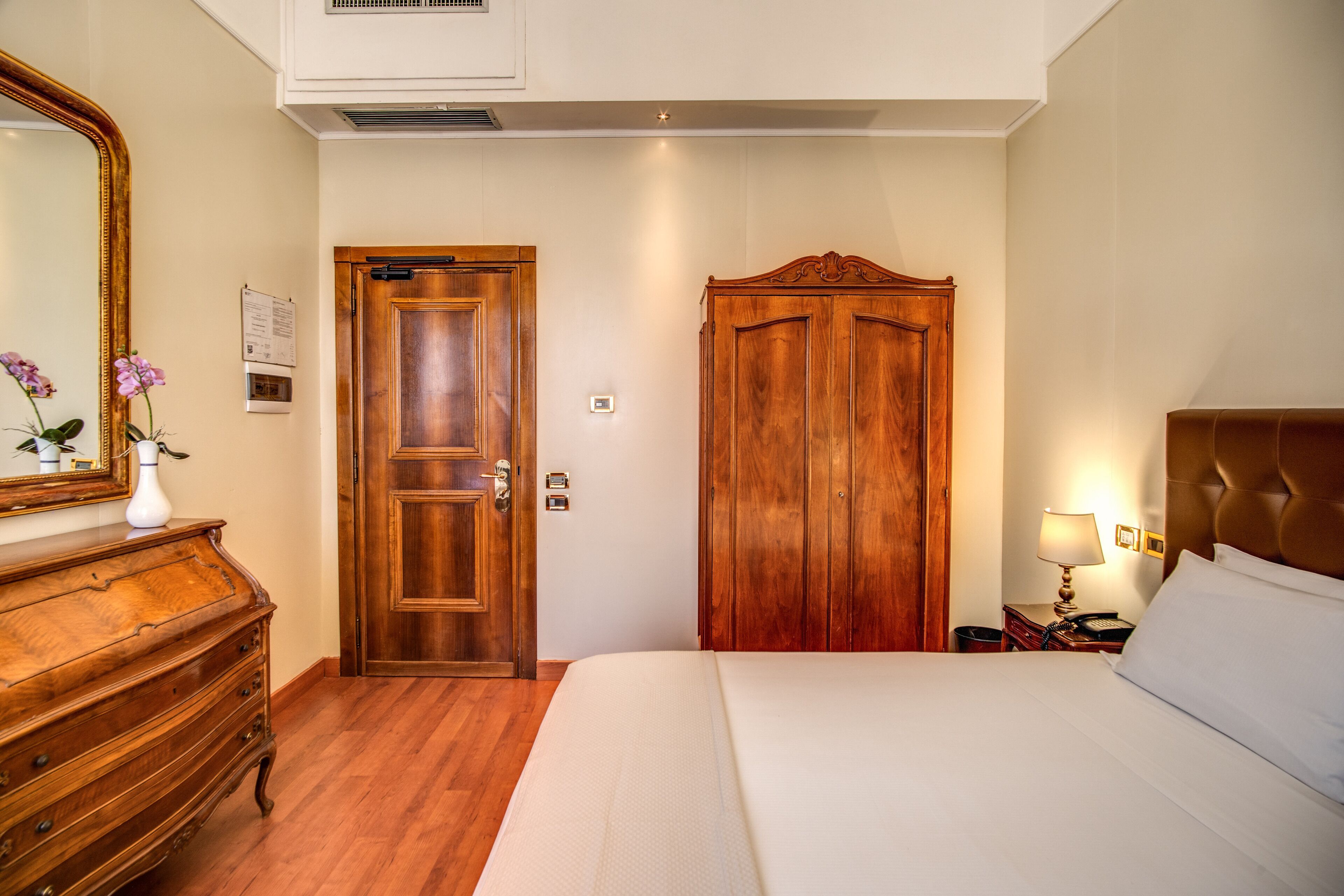 A hotel room with a wooden dresser, a large bed, a wardrobe, and a nightstand with a lamp.