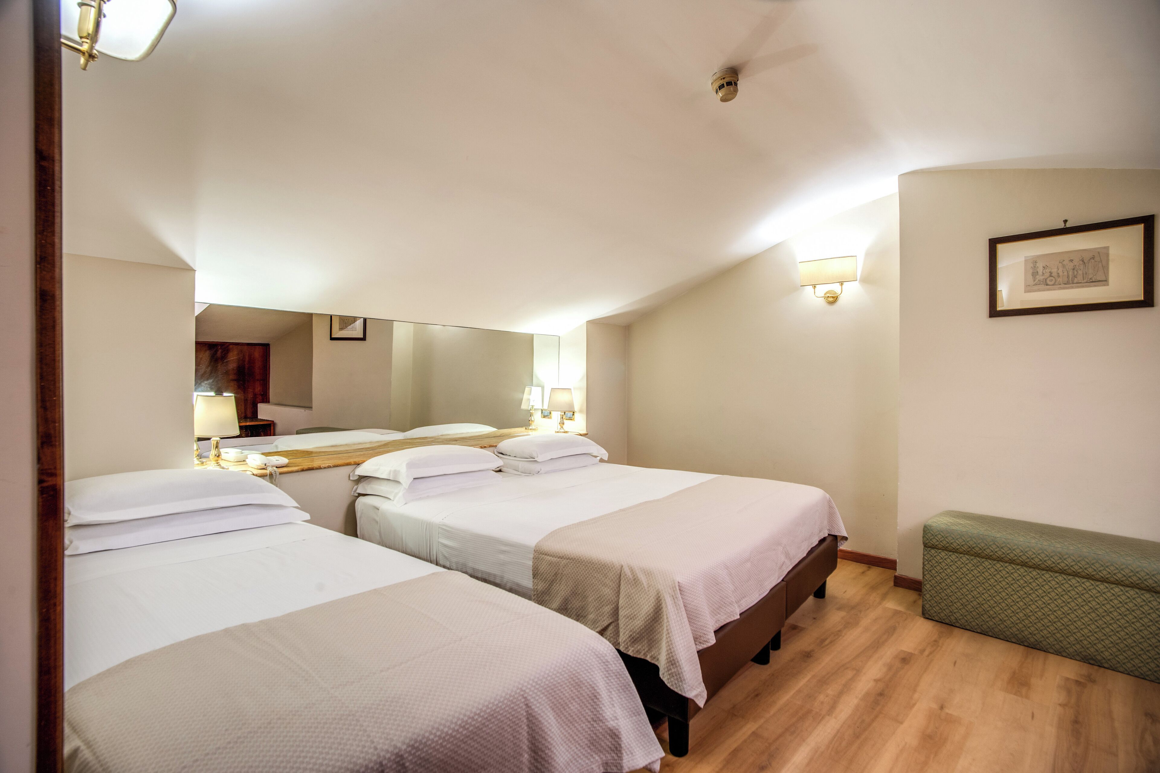 A hotel room with three single beds, a bench, a mirror, and a framed picture on the wall.