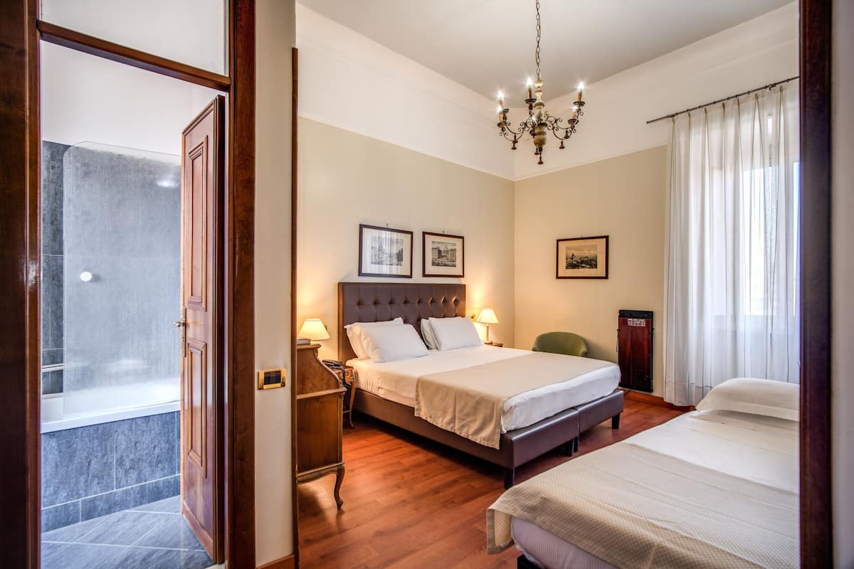 A hotel room with a large bed, a smaller bed, a vanity with a mirror, a chandelier, and framed pictures on the wall.