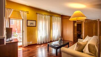 Una sala de estar con sofá, una mesa de centro y televisión. Hay cortinas en las ventanas y una lámpara sobre la mesa.