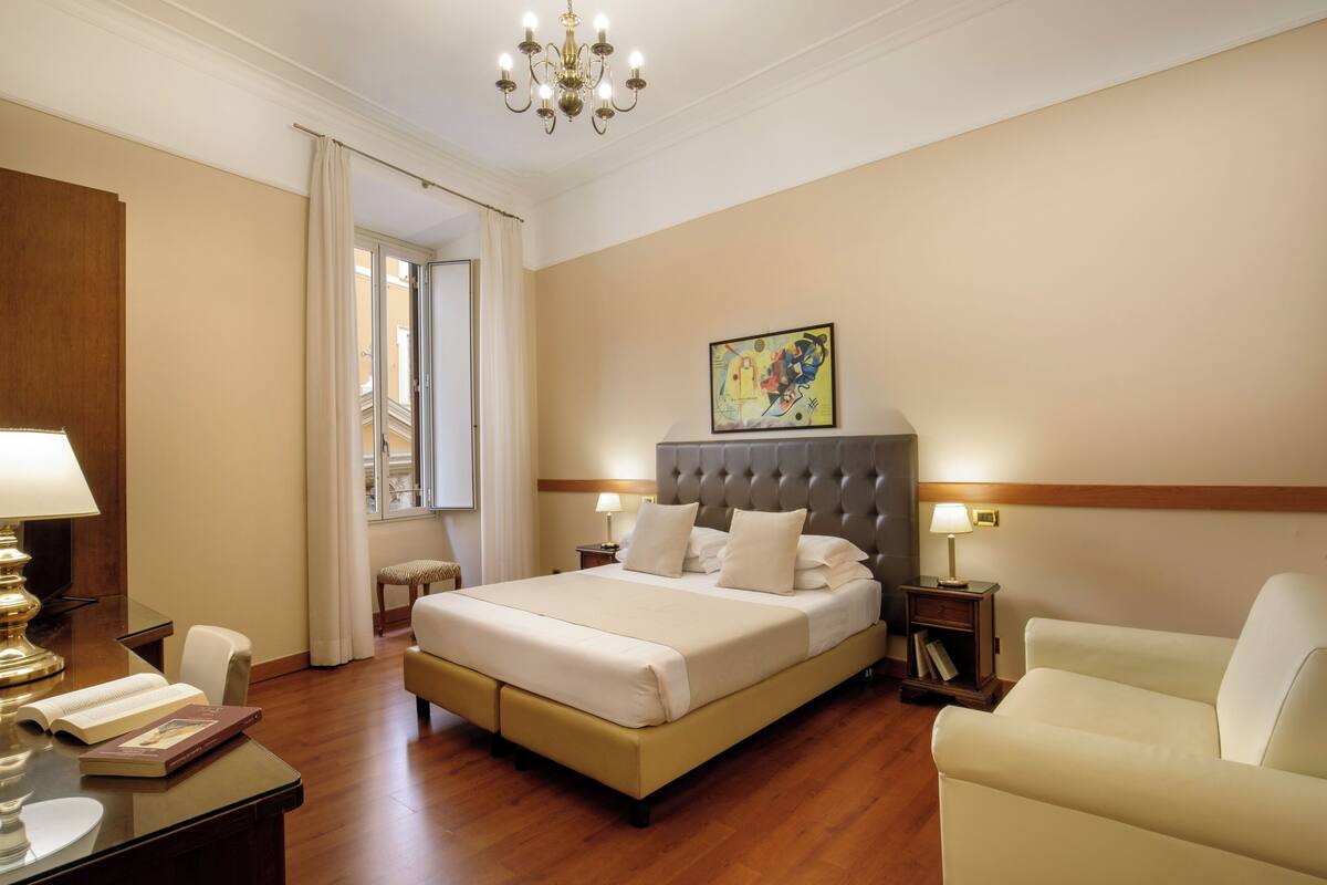 A hotel room with a large bed, a desk with a lamp, a chair, and a nightstand with a lamp.