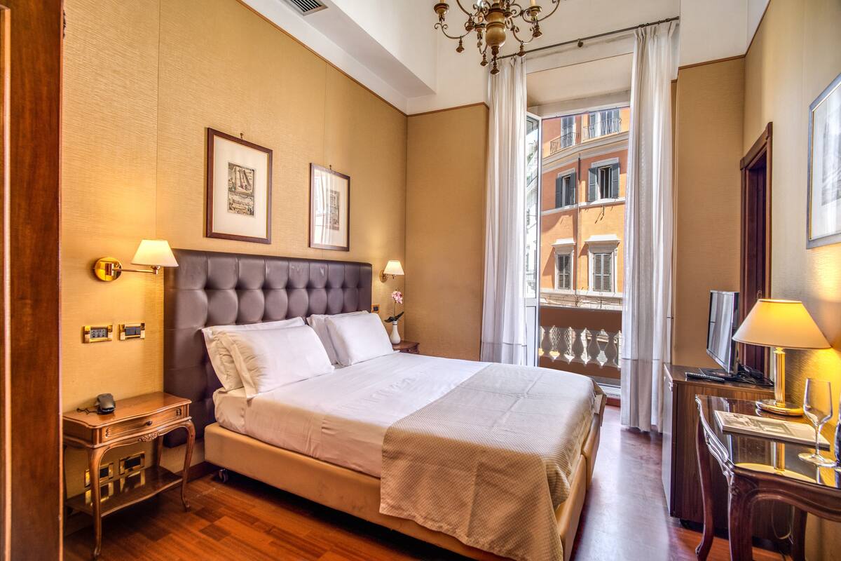 A hotel room with a large bed, a desk, a chair, a TV, and a balcony with a view of buildings.