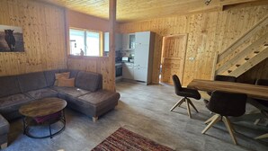 Ein gemütliches Blockhaus-Interieur mit einer Eckcouch, einem kleinen runden Tisch, einem Esstisch mit Stühlen und einer Küchenzeile.