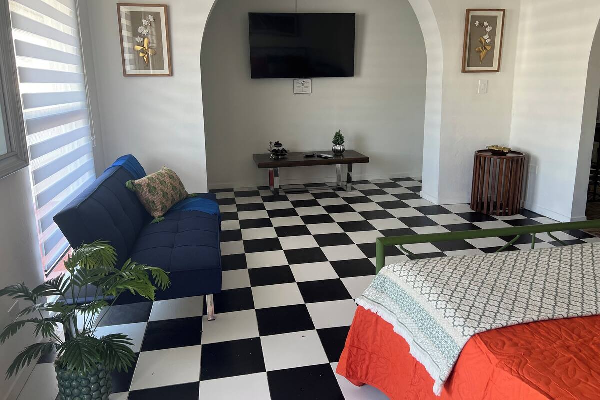 A room with a checkered floor, a blue sofa with a plant, a bed with a patterned bedspread, a small table with a plant, and a TV mounted on the wall.