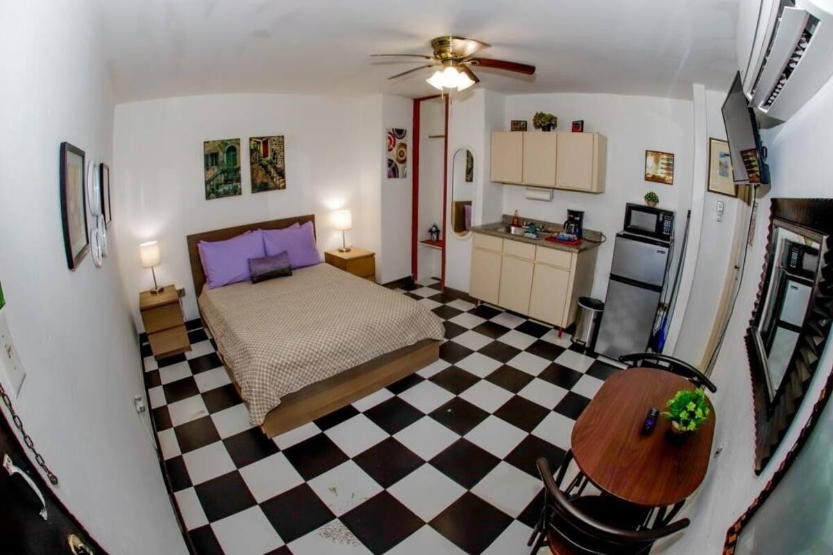 A compact room with a bed, a small table, a ceiling fan, a kitchenette, and a checkered floor.