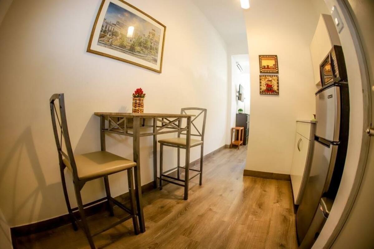 A compact kitchen with a dining table and chairs, a refrigerator, and a framed picture on the wall.
