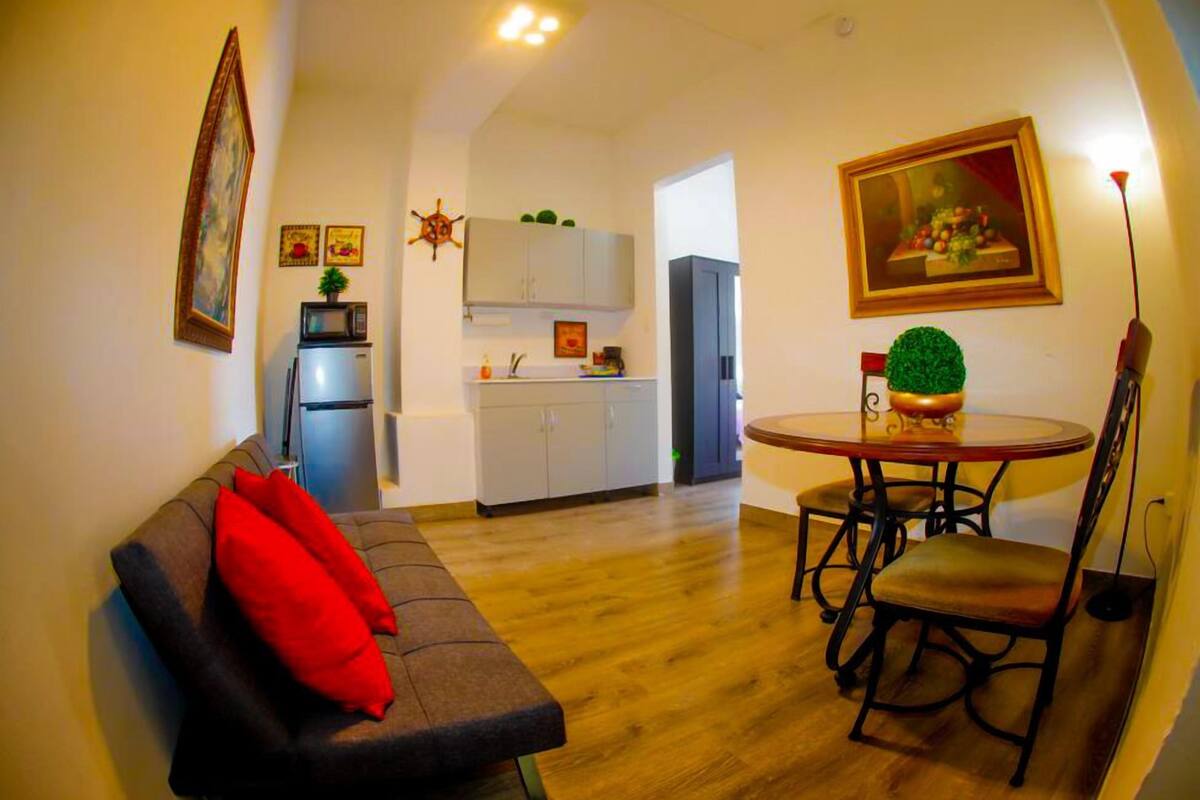 A compact living space with a sofa, a dining table with chairs, a kitchen area, and a small dining table with a bowl.