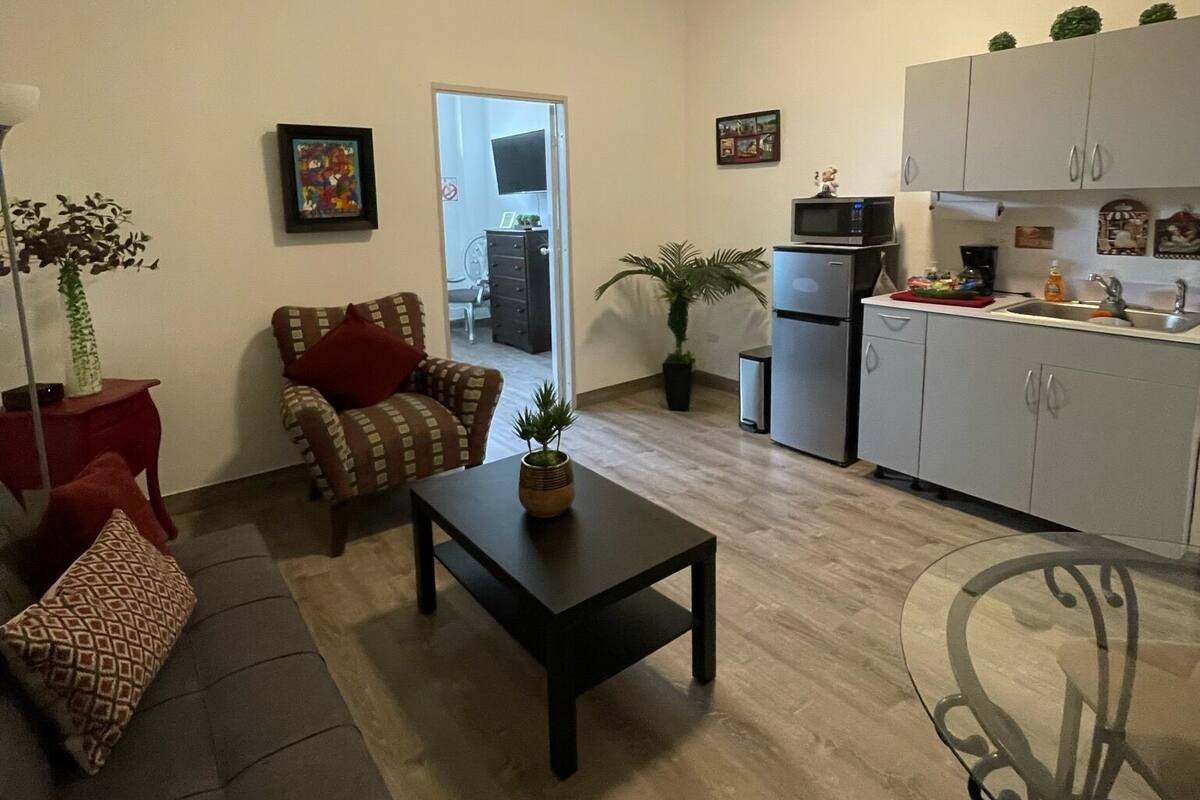 A compact living space with a sofa, a coffee table, a dining table, a kitchen area with cabinets and appliances, and a door leading to another room.