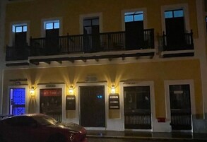 Un edificio de dos pisos con un balcón en el nivel superior, un cartel y un coche estacionado frente a él.