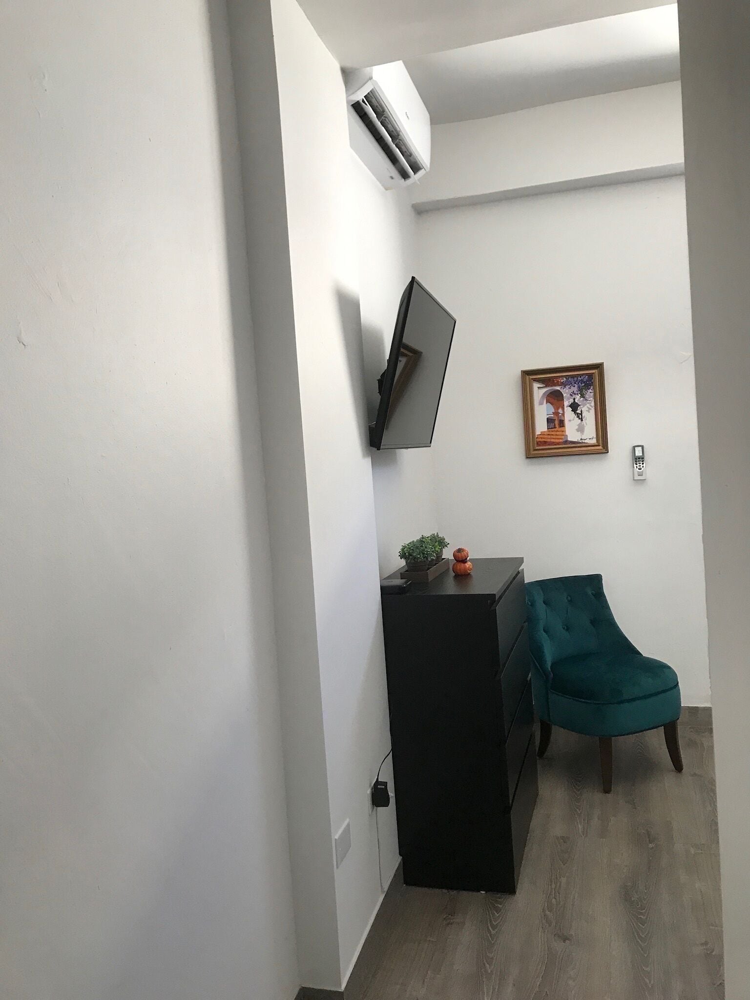 A small, modern room with a flat-screen TV mounted on the wall, a small table with a plant, a teal chair, and a framed picture.