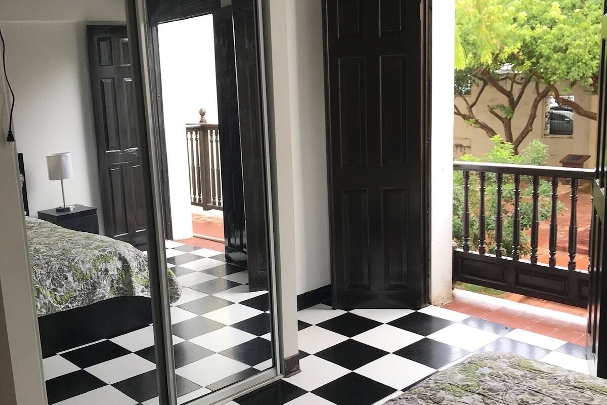 A room with a checkered floor, a bed with a floral bedspread, a mirror, and a door leading to an outdoor balcony with a view of greenery.