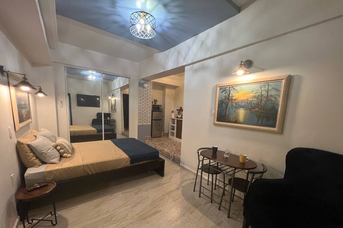 A modern bedroom with a bed, a small table, a chair, and a painting on the wall.