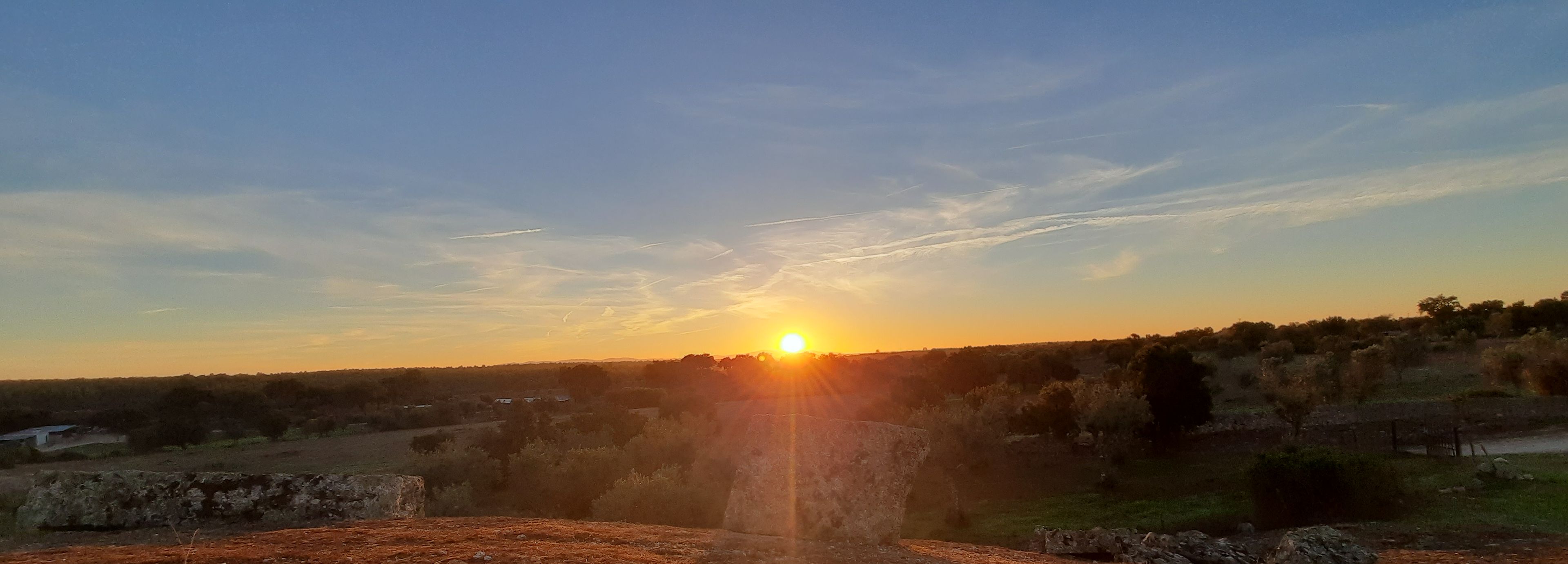 Un coucher de soleil sur un paysage rural, avec un chemin de terre menant à une construction en pierre.