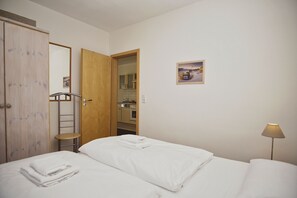 Une chambre d’hôtel avec un lit, une armoire en bois, un petit tabouret, une porte donnant sur une cuisine et un tableau accroché au mur.