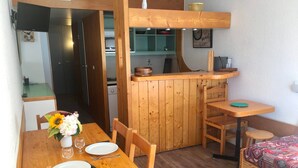 A compact wooden room with a dining table set for two, a small kitchen area, and a bed with patterned bedding.