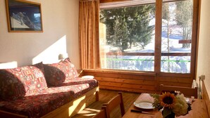 A cozy room with a wooden sofa, a dining table with a plate and a vase of flowers, and a window with a view of trees and snow.