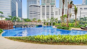A swimming pool with a bridge, surrounded by greenery and palm trees, with tall buildings in the background. - EBONY by Kozystay (Jakarta)