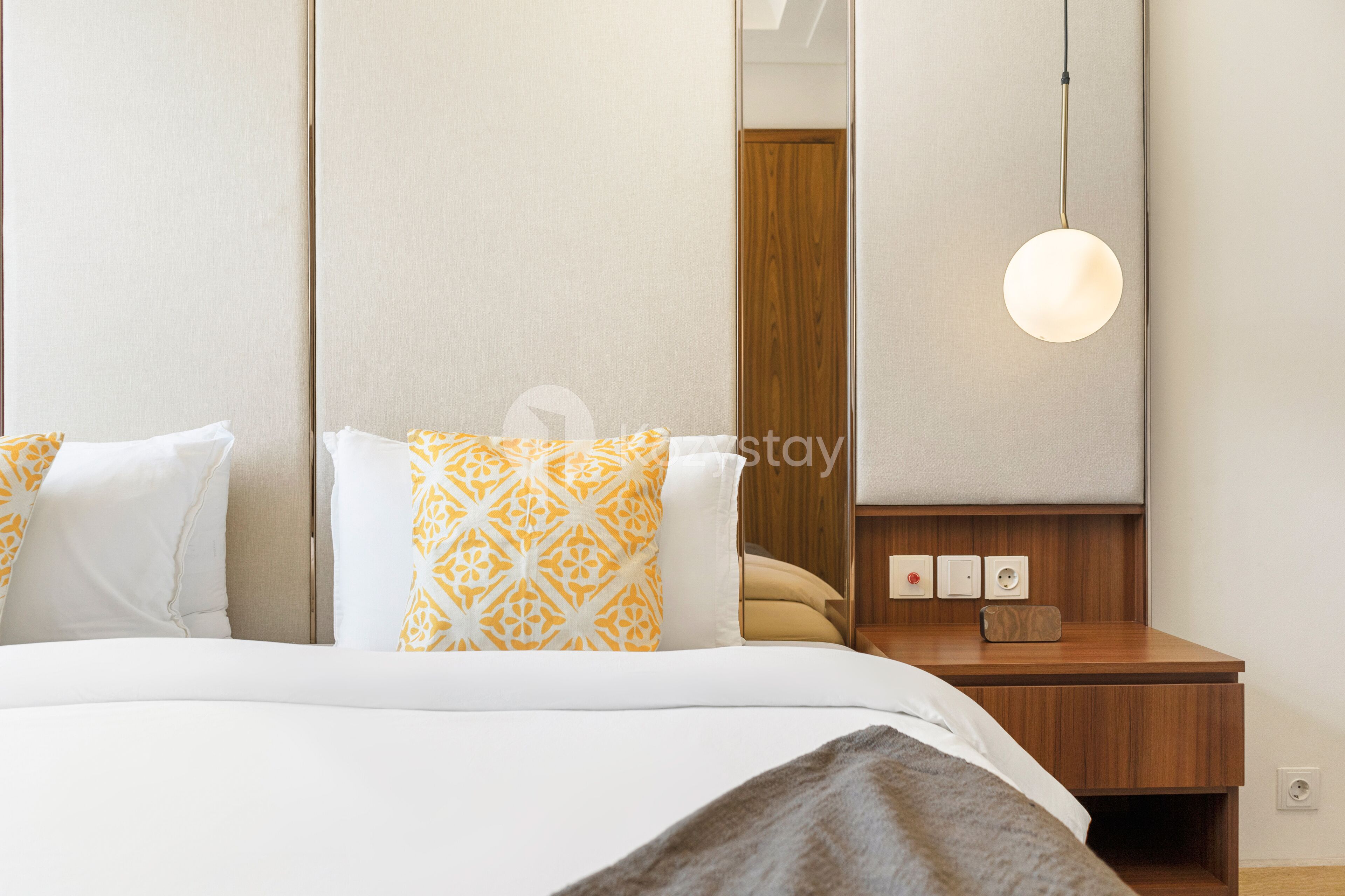 A hotel room with a bed, a bedside table, a mirror, and a wall-mounted light fixture.