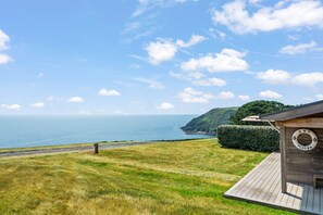 Een landhuis aan de kust met een houten terras, een reddingsboei en uitzicht op zee en heuvels.