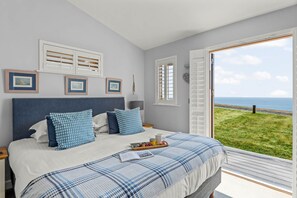 A bedroom with a bed, a nightstand, a window with a view of the sea, and a door leading outside. - House 38 - sleeps 6 guests in 3 bedrooms (Looe)