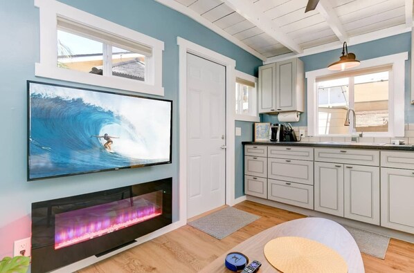 A modern kitchen with a flat-screen TV mounted above a fireplace, light-colored cabinets, and a window providing natural light.