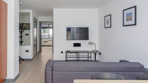 Un salon moderne avec un canapé gris, un téléviseur à écran plat fixé au mur et une petite table avec une lampe.