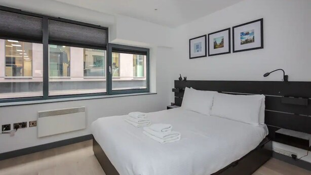 Une chambre d’hôtel moderne avec un grand lit, une fenêtre avec des stores, un radiateur et trois tableaux encadrés accrochés au mur.