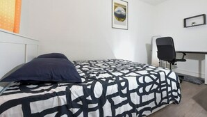 Een enkel bed met een geborduurd beddekleed, een zwarte stoel, een bureau en een ingelijst beeld aan de muur.