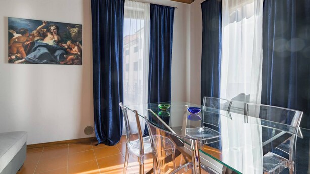 A room with a glass dining table, clear chairs, a painting on the wall, and blue curtains. - SUITE ARENA IN CENTRO CITTA' (Verona)