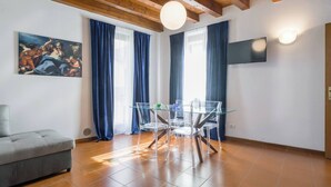 A living room with a sofa, a glass dining table with chairs, a painting on the wall, and a TV mounted on the wall. - SUITE ARENA IN CENTRO CITTA' (Verona)
