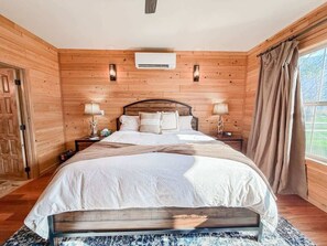 A wooden-paneled bedroom with a bed, bedside lamps, an air conditioning unit, a bench, and a window with curtains.