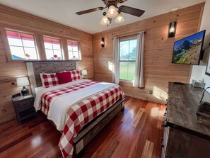 A bedroom with a wooden floor, a bed with a checkered bedspread, a nightstand, a ceiling fan, and a window with a mountain view.