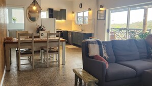 Un salón amplio con un sofá gris oscuro, un banco de madera y una alfombra estampada. Una mesa de comedor con sillas está situada en el centro, y se ve una cocina con ventana al fondo.