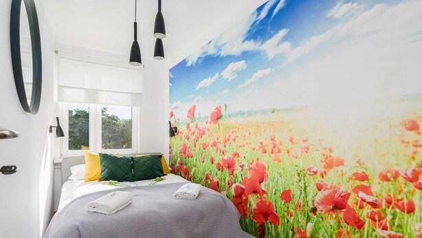 A bedroom with a large window, a bed with a grey quilt, green and yellow pillows, and a wall mural of red poppies.