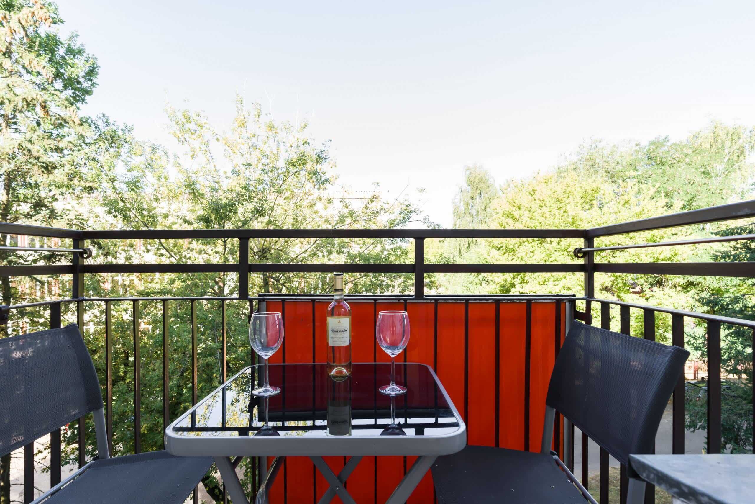 Un tavolo con due bicchieri da vino e una bottiglia su un balcone con ringhiera in metallo e sfondo rosso.