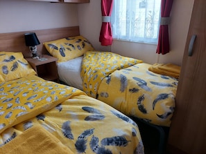 A room with two beds, each with yellow bedding featuring a feather pattern. There is a nightstand with a lamp and a small table with a book.