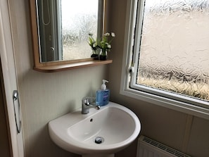 A bathroom with a white sink, a mirror, a small shelf with a vase of flowers, and a window with frosted glass.