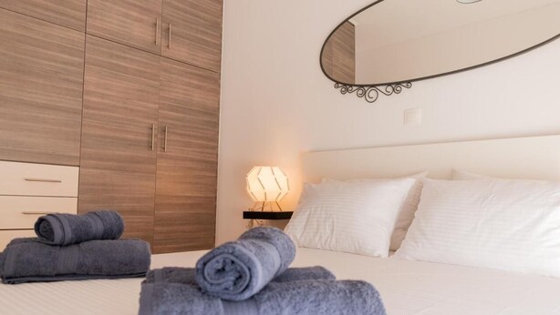 A neatly made bed with white linens and rolled gray towels. There is a bedside lamp with a geometric light fixture. A large mirror with a curved frame is mounted on the wall above the bed. - GTD31502 - Art room of Athens - Hera (Palaio Faliro)