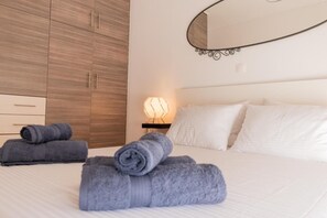 A neatly made bed with white linens and rolled gray towels. There is a bedside lamp with a geometric light fixture. A large mirror with a curved frame is mounted on the wall above the bed.