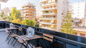 A rooftop terrace with wooden tables and chairs, a white lantern, and a view of multi-story buildings and trees. - GTD31502 - Art room of Athens - Hera (Palaio Faliro)