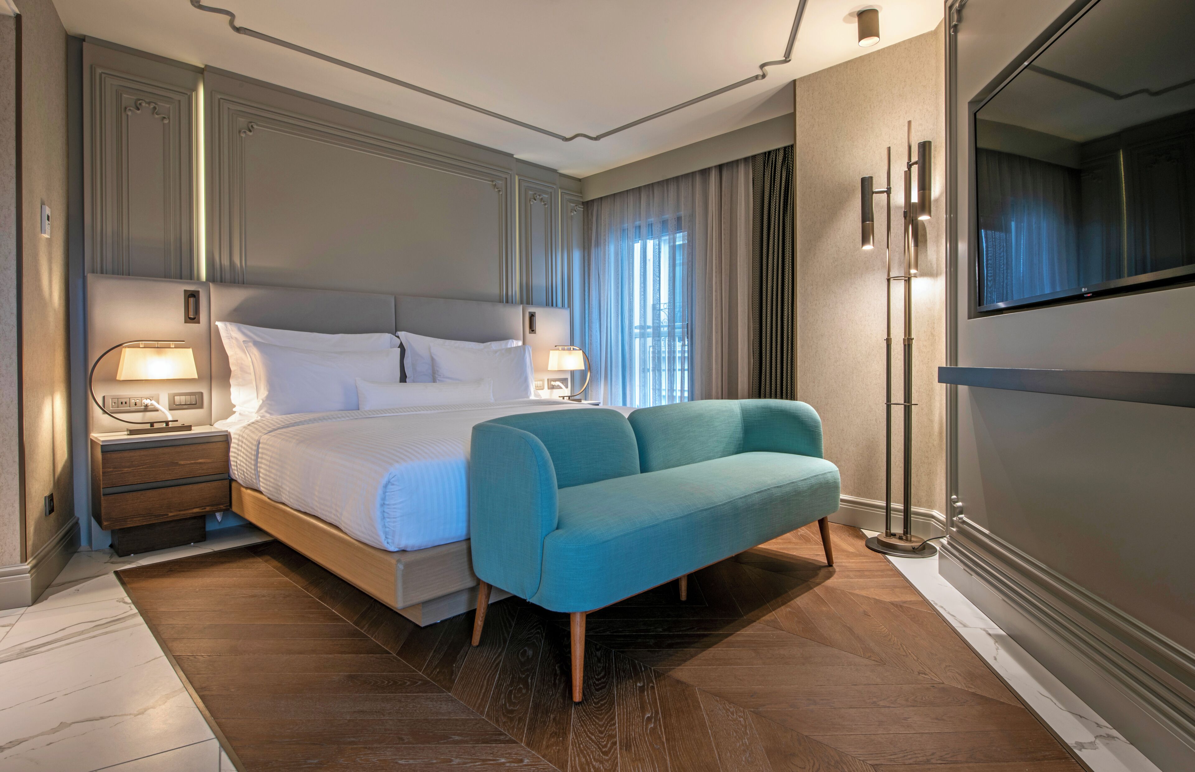 A modern hotel room with a large bed, a sofa, a nightstand with lamps, and a large mirror.