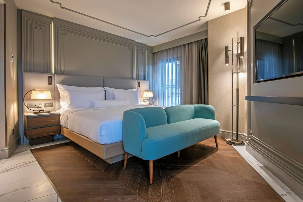A modern hotel room with a large bed, a sofa, a nightstand with lamps, and a large mirror.