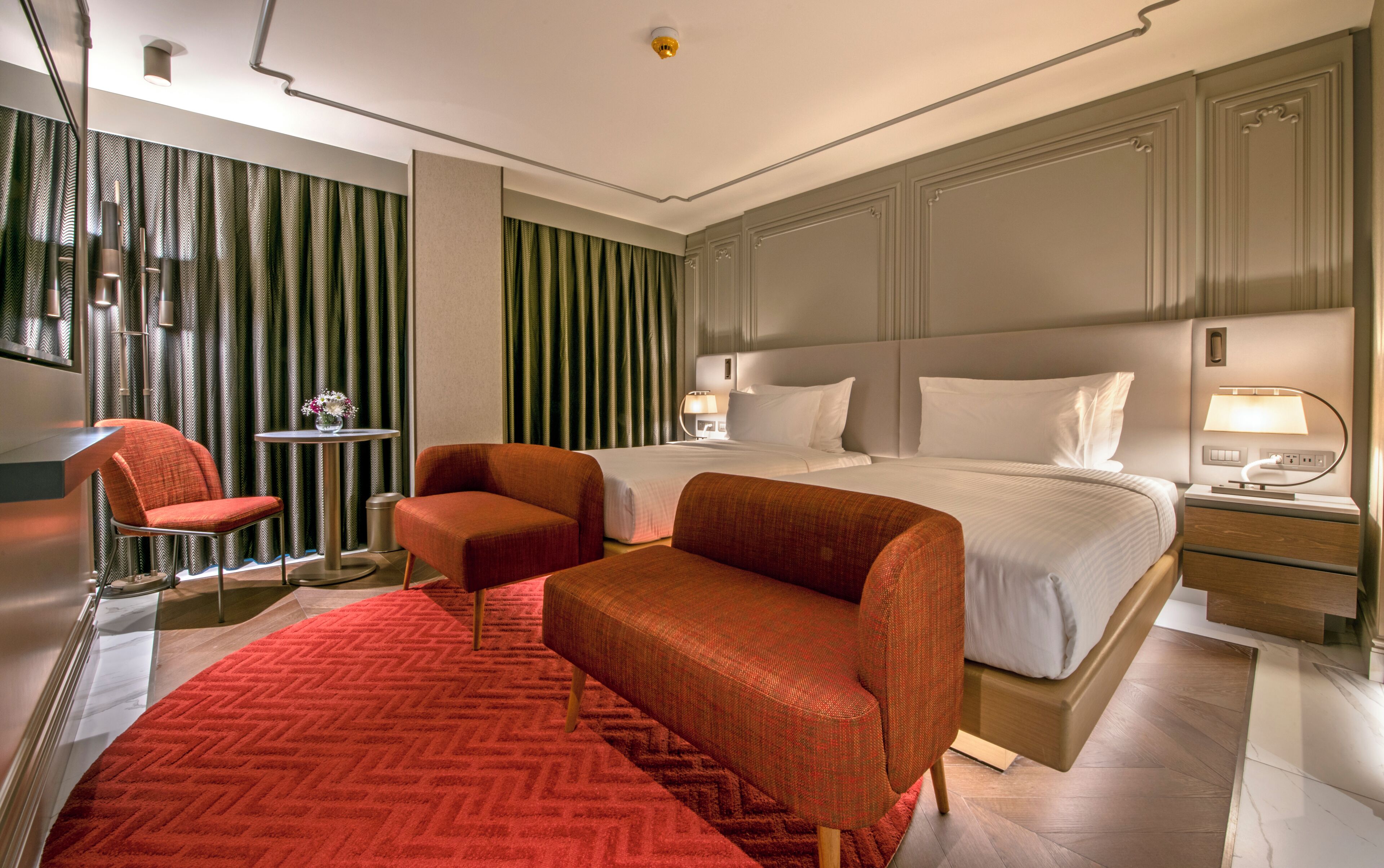 A hotel room with a large bed, two armchairs, a small table with a vase, and a red carpet.